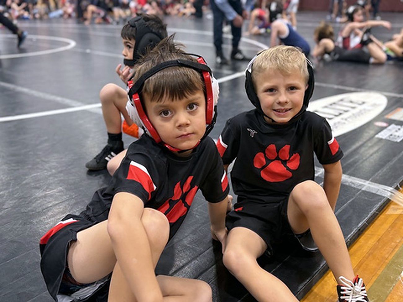 Two young MSO wrestlers in headgear sitting on the mat between matches.